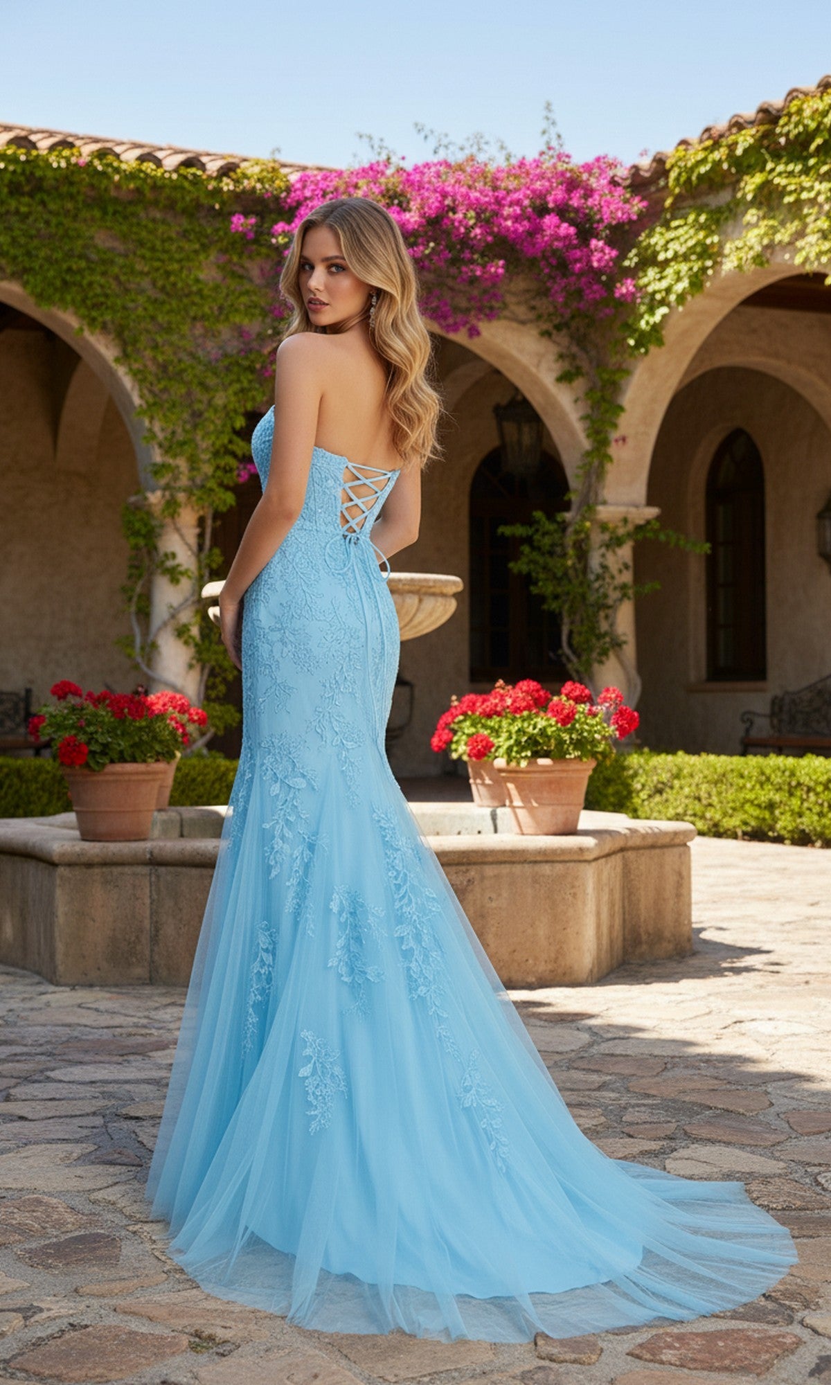 Wearing the PromGirl Exclusive 250227, a woman in a light blue, strapless lace prom dress with a lace-up back stands outside by potted flowers and a stone building draped in greenery.